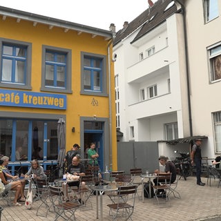 Vor dem "Café Kreuzweg" sitzen auf Klappstühlen mehrere Menschen an einem Tisch und essen.
