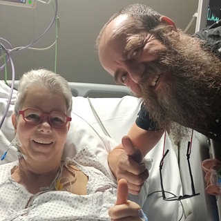 Eine Frau liegt in einem Krankenhausbett, daneben steht ein Mann mit Vollbart. Beide strecken ihre Daumen nach oben. 