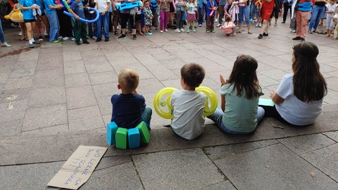 Vier Kinder sitzen auf dem Neuenburger Rathausplatz. Im Hintergrund stehen die Demonstrierenden. 