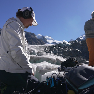 Ein Mann kniet im Schnee auf einem Gletscher und nimmt Geräusche auf. Im Hintergrund spielt ein Mann auf einer Klarinette.