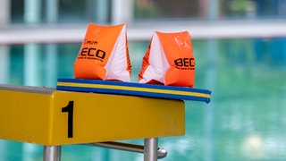 Zwei Schwimmflügel und ein Schwimmbrett liegen auf einem Startblock in einer Schwimmhalle.