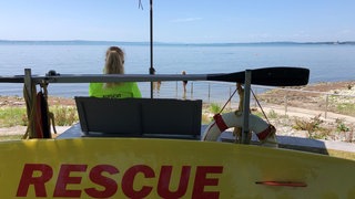 Eine Frau sitzt am Bodenseeufer unter einem Sonnenschirm. Die DLRG am Bodensee bereitet sich auf das erste Hitzewochenende in der Region vor.