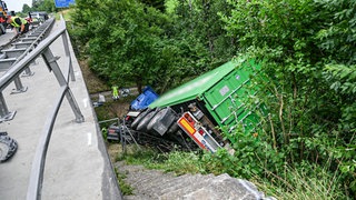 Einsatzkräfte arbeiten an der Unfallstelle. Ein Lastwagen hat auf der A96 bei Wangen im Allgäu die Leitplanke durchbrochen und neben einer Autobahnbrücke liegt im abschüssigen Gelände. 