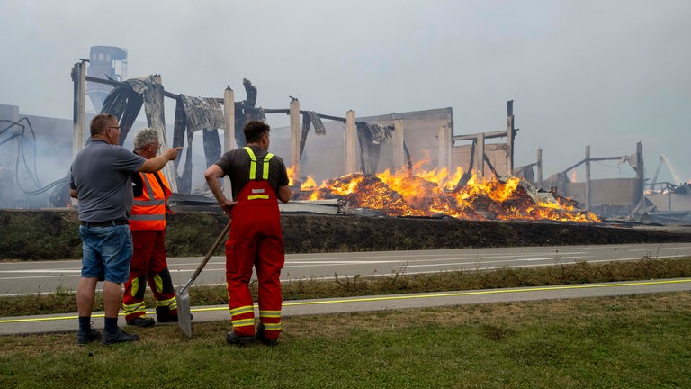 Zwei Feuerwehrleute unterhalten sich mit einem Mann vor den Trümmern eines brennenden holzverarbeitenden Betriebs