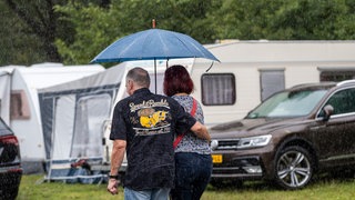 Ein Paar mit Regenschirm auf einem Campingplatz (Symbolbild). Wie sieht es bei dem Dauerregen mit Camping in der Region aus? Wir haben auf den Campingplätzen nachgefragt. 