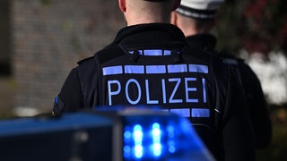 Zwei Polizeibeamte, von hinten zu sehen, stehen hinter einem Polizeiauto. Im Vordergrund ist noch das Blaulicht des Fahrzeugs zu sehen. Mehrere Männer sollen der Polizei zufolge in Bad Mergentheim (Main-Tauber-Kreis) mit einer Machete und einer Schreckschusswaffe aufeinander losgegangen sein. (Symbolbild)