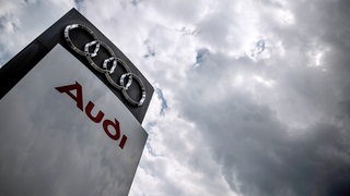 Dunkle Wolken über einen Audi-Logo (Symbolbild). Gerüchte um Kapazitätskürzungen und Sparpläne sorgen für Unruhe bei Audi. Besonders das Werk Neckarsulm ist in den Fokus gerückt. 