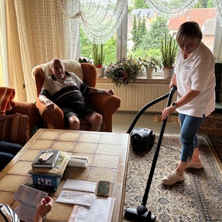 Eine Haushaltshilfe saugt in einem Wohnzimmer den Teppich, während ein älterer Mann auf einem Sessel sitzt, eine ältere Frau auf einem Sofa und eine jüngere Frau sich Briefe anschaut.