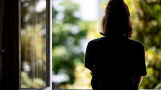 (Symbolbild) Eine Frau steht in ihrer Wohnung an einem Fenster.