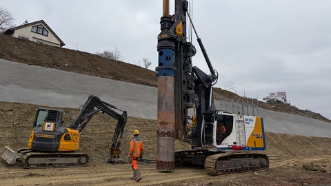 Ein Bauarbeiter und ein Bagger stehen neben einer sehr großen Bohrmaschine, die Löcher in den Boden der neuen A8 Strecke bei Pforzheim bohrt.