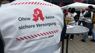 Apotheker aus Karlsruhe stehen an Stehtischen vor einem Einkaufszentrum in Karlsruhe. Sie tragen weiße Westen mit der Aufschrift "Ohne Apotheken keine Versorgung". 