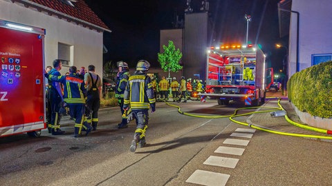 Feuerwehrmänner stehen nachts im Licht von Scheinwerfern vor Feuerwehrautos und einem Lagerhaus 