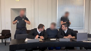 Der Angeklagte sitzt zwischen seinen Verteidigern auf der Anklagebank, dahinter zwei Justizbeamte
