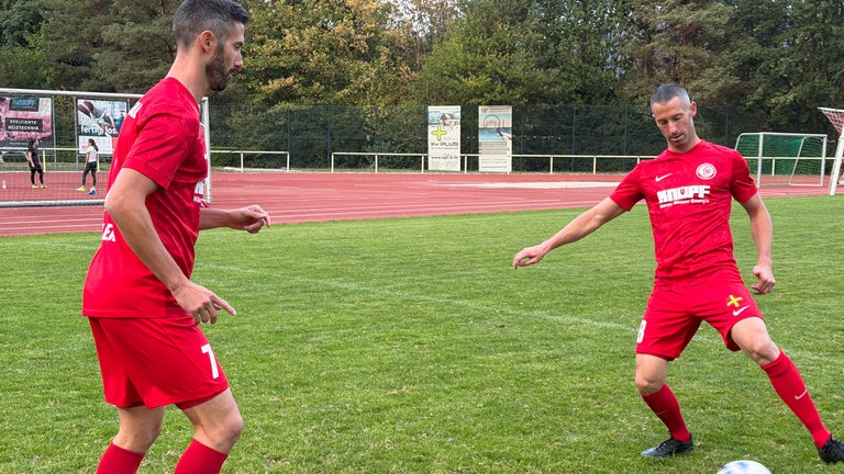 Zwei Zwillinge in rotem Trikot passen sich ein Fußball zu.