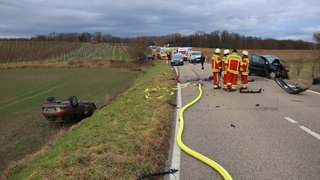 Ein Auto ist drei Meter eine Böschung herunter gestürzt und liegt auf dem Dach. Auf der Fahrbahn liegen Autoteile. Im Hintergrund stehen Krankenwagen.