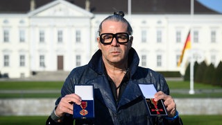 Fotograf Luigi Toscano steht vor dem Schloss Bellevue und hält in der Hand Verdienstmedaille und Verdienstkreuz. 