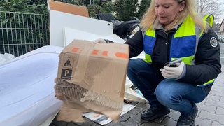 Sabrina Brand kniet neben einem Sperrmüllhaufen. Mit Handschuhen und Warnweste sucht sie nach Namen und Adressen.