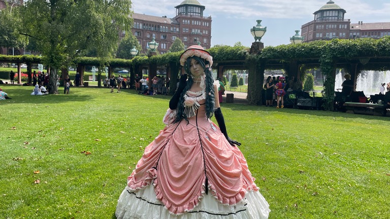 Zum Outfit gehören eine blaue Perücke, ein mit Rosen besetzter Hut und ein langes, rosafarbenes Kleid mit Rüschen
