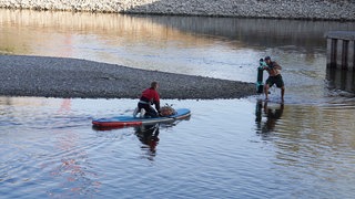 Ein Umweltschützer zieht einen E-Scooter bei Niedrigwasser aus dem Neckar. Eine weitere Freiwillige kommt mit dem Stand Up Paddle-Board angefahren, um den Roller aufzuladen.