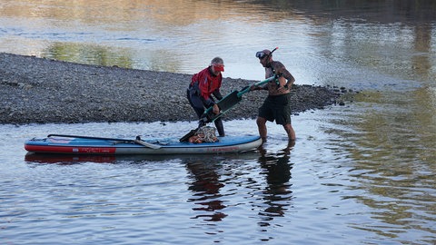 Umweltschützer Uwe Franken zieht einen E-Scooter bei Niedrigwasser aus dem Neckar. Eine weitere Freiwillige kommt mit dem Stand-Up-Paddle-Board angefahren, um den Roller aufzuladen.