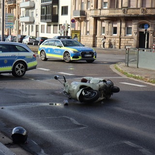 Auf der Straße liegt ein Roller auf der Seite. Davor liegt ein Helm. Hinter dem Auto sieht man zwei Polizeiautos, die die Straße absperren.