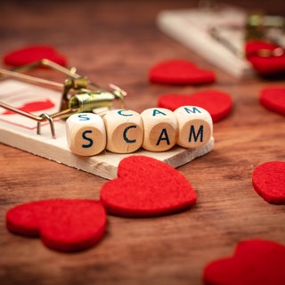 Eine Mausefalle, rote Herzen und Würfel mit Buchstaben, die das Wort "Scam" bilden (Symbolbild "Love Scamming").