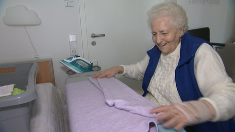 Die 90-jährige Sofia Thut sitzt vor ihrem Bügelbrett und faltet ein Kleidungsstück.