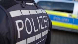 Das Logo der Polizei auf der Dienstkleidung eines Polizisten. Im Hintergrund ein Polizeiauto. 