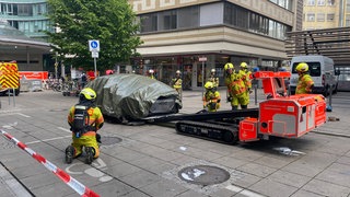 Feuerwehrleute transportieren ein abgedecktes E-Auto nach einem Brand in einer Tiefgarage mit einem speziellen Gerät ab.