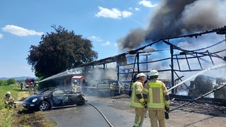 Vor einem abgebrannten Gebäude stehen zwei Feuerwehrmänner. Aus dem Gebäude raucht es. Davor ein ausgebranntes Auto.