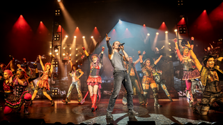 Das Ensemble des Musicals "We will rock you" steht in bunten Kostümen auf der Bühne in Stuttgart und tanzt.