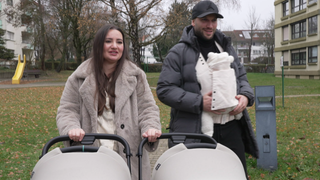 Mutter und Vater gehen mit zwei Kindern im Kinderwagen und einem Kind in der Trage spazieren.