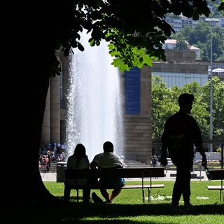 Menschen sitzen im Schlossgarten bei sommerlichen Temperaturen und strahlendem Sonnenschein im Schatten unter einem Baum auf einer Bank. 