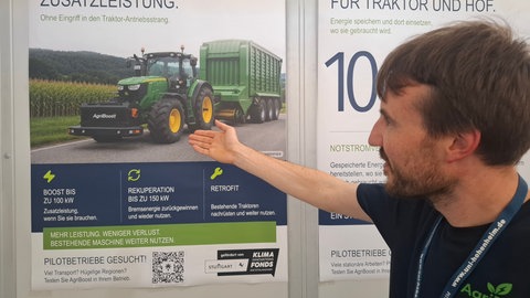 Der "AgriBoost" vorne am Traktor: Philipp Seitz zeigt auf einem KI-generierten Bild, wie das Gerät künftig eingesetzt werden könnte. Da bei schweren Landmaschinen ohnehin ein Ausgleichsgewicht vorne am Traktor für Fahrsicherheit und Traktion notwendig ist, dient die E-Unterstützung zudem als Frontgewicht.  