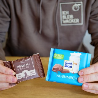 Matteo Wacker, Inhaber und Firmengründer der Wacker GmbH, hält eine Tafel mit Schokolade von Ritter Sport und einen Haferriegel von Wacker mit der Aufschrift "Monnemer Quadrat Bio" in den Händen. (Archivbild)