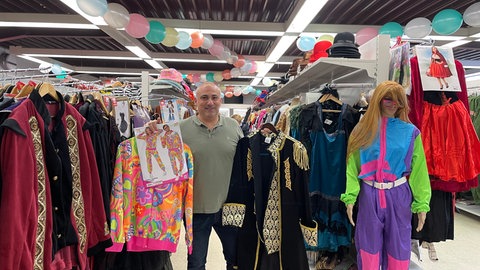Alexandros Theodosiadis steht im Gang voller Kostüme und präsentiert zwei Trendoutfits dieser Faschingssaison. Ein Hippiekostüm und ein Piratenkostüm.