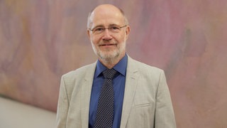 Harald Lesch steht in Anzug und Krawatte vor einer pastellfarbenen Wand im Schloss Bellevue in Beriln