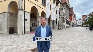 Der OB von Albstadt, Roland Tralmer (CDU) hält ein Autokennzeichen mit der Aufschrift "ALB- IN- 26" in den Händen.