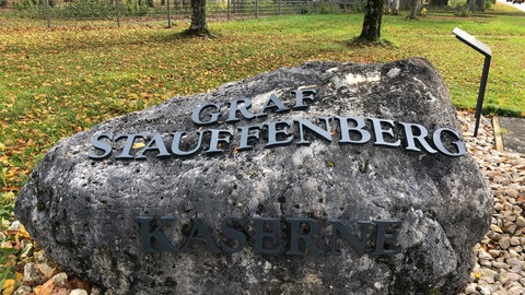 Auf einem Stein steht "Graf Stauffenberg Kaserne", im Hintergrund sind Kasernengebäude zu sehen. Das Bundesverteidigungsministerium plant, ehemalige Bundeswehrliegenschaften zu behalten.
