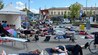 Schweigend liegen mehrere hundert Menschen auf dem Boden. Auf Feldbetten, Isomatten und Piknikdecken machen sie auf die Krankheit MECFS aufmerksam