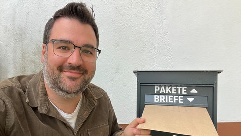 Andreas Hummel schaut in die Kamera, während er einen Brief in den Briefkasten eines öffentliches Gebäudes wirft. Er ist Bürgermeister-Kanddidat in Egesheim