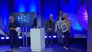 Die Beteiligten rund um ein Stehpult auf der Bühne, links die Moderatorin, rechts Intendant und Geehrte. Die Träger des Hans Bausch Mediapreises halten ihre Urkunden in die Kameras.