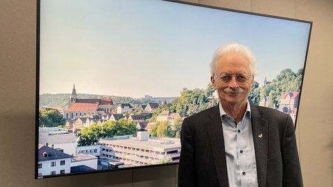 Vor einem Bildschirm, der eine Stadtansicht von Tübingen zeigt, blickt Hans-Georg Rammensee freundlich in die Kamera. Der Immunologe forscht auch mit über 70 Jahren weiter zu Krebstherapien. 