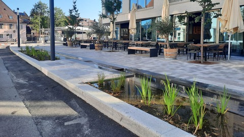 Blick auf das Drei-Kronen-Quartier in Tuttlingen mit flachem Wasserbecken, schmalem Wasserlauf, Sitzbereichen und Bäumen zwischen Wohn- und Geschäftsbauten, die zusammen für ein kühleres Stadtklima sorgen.