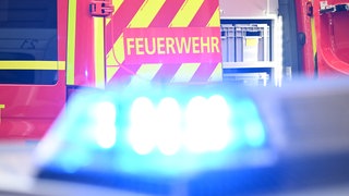 Ein Fahrzeug der Feuerwehr steht bei einem Einsatz auf einer Straße, während im Vordergrund das Blaulicht eines Polizeifahrzeugs zu sehen ist.