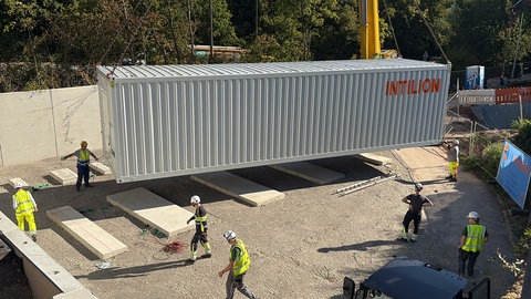 Ein großer grauer Container wird mit einem Schwerlastkran auf eine Betonfläche gesetzt. Das ist der neue Batteriespeicher der Uniklinik Tübingen.