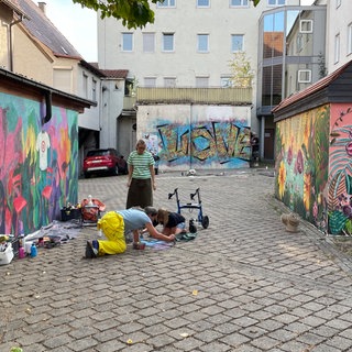 Ein Hof zwischen Wohnhäusern. Rechts und links sind Garagen bunt bemalt, auf der gegenüberliegenden Wand kann man groß "Love" lesen. In der Mitte beugen sich drei Personen über Pflastersteine, die sie gerade bemalen. 