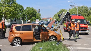 Ein orangefabenes Auto steht an einer Kreuzung, umgeben von Rettungskräften. Vor dem Auto ist ein umgekippter Ampelmast zu sehen. Er hat eine Fußgängerin bei einem Autounfall schwer verletzt.