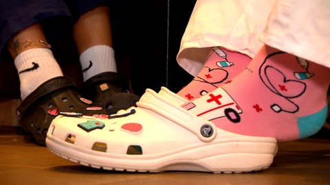 Im Vordergrund sind weiße Schuhe mit Riemen und Löchern und bunten, aufgesteckten Figuren wie ein Gehirn oder ein OP-Kittel zu sehen, darin Stecken Füße in pinken Socken mit Krankenwagen und Stethoskop. Im Hintergrund sind ähnliche Schuhe in schwarz zu sehne. Die Schwestern Lily und Sina vereint die Passion für den Pflegeberuf.