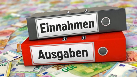 Zwei Aktenordner mit der Aufschrift "Einnahmen" und "Ausgaben" liegen auf Geldscheinen (Symbolfoto): Hunderte Kommunen in Baden-Württemberg haben massive Rückstände bei ihren Jahresabschlüssen. 